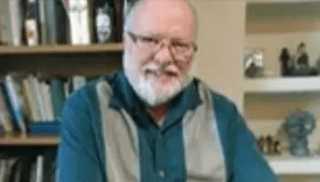 An Older Man With a Beard and Glasses Is Seated at a Table, Looking Thoughtfully at Something in Front of Him.